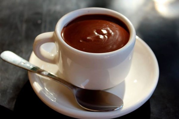 A white cup of thick, rich dark brown drinking chocolate on a white saucer with a spoon.