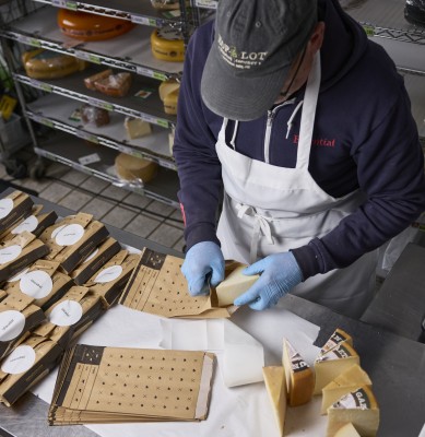 An overhead shot of a table in the cheese room with labeled bags of cheese on the left and fresh cut wedges of cheese on the right. A worker in the middle is placing a wedge of cheese into a bag. In the background are whole wheels of cheese.