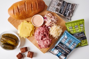 The contents of a Reuben kit laid out on a board, including rye bread, a mound of sliced deli meat, sauerkraut, coleslaw, swiss cheese, brownie bites, pickles, bags of potato chips, and a ramekin of russian dressing.