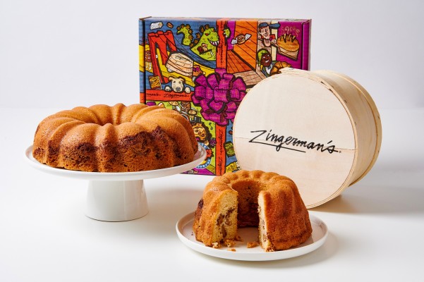 A photo of a small nosher sized coffee cake on a plate and a large fresser coffee cake on a cake stand, both in front of a cartoon gift box and a wooden crate.